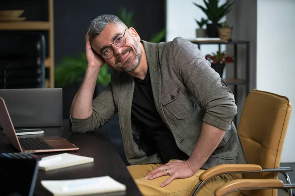 stock image Happy worker sits in loft office, smiling with contentment work. Desk with computer, stationary, and other necessary tools, reflecting productive workplace where technology and comfort merge