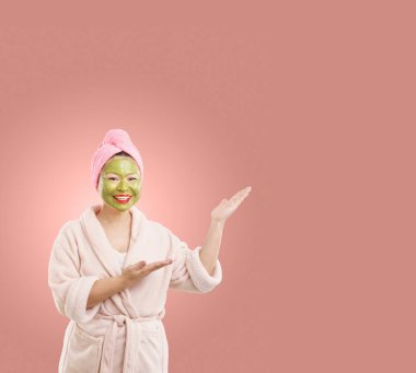 Yüzüne yeşil kozmetik maske takan orta yaşlı kadın, izole toz rengi arkaplanındaki boş fotokopi alanına işaret ederek temiz ve taze estetik kadın güzelliği ve cilt bakımını ekliyor. Yüksek