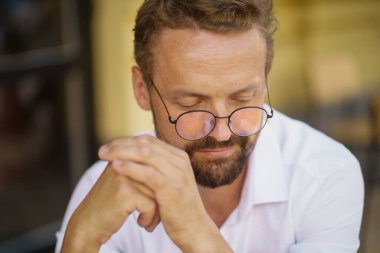 Yumuşak görünüşlü, gözlüklü beyaz bir adam. Avrupa 'nın büyüleyici caddesinde oturuyor. Onun şık görünüşü modern ve sıradan modanın karışımını yansıtıyor, kendine güven ve zeka yayıyor.