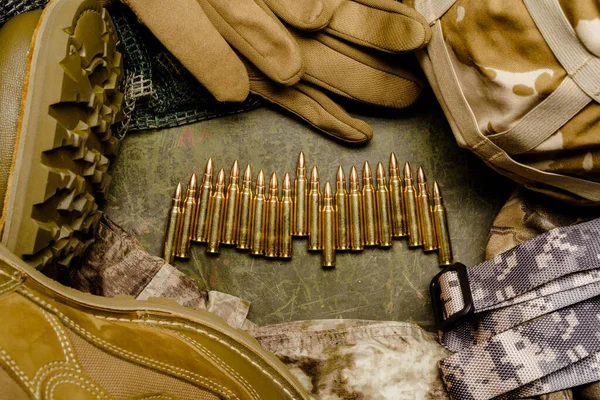 Set of nails for 5,56 in center, helmet, military boots, gloves placed next to a bunch of bullet casings. The gloves are resting on a textured surface.