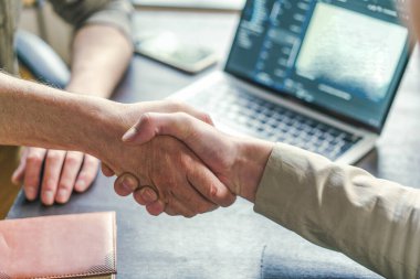 Bir dizüstü bilgisayarın önünde el sıkışan iki kişiyi gösteren bir fotoğraf. İş, takım çalışması ve profesyonel bir ortamda işbirliği simgeliyor..