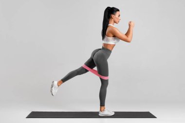 Gri arka planda direnç bandıyla kalça kasları için egzersiz yapan atletik kız. Spor salonundaki kadın.