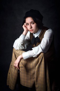 Genç bir kadın dedektifin karamsar stüdyo portresi. Klasik Viktorya tarzı kıyafetler giyiyor, ekose ceket ve beyaz gömlek giyiyor. Sherlock Holmes 'un kamusal alandaki ikonik figüründen esinlenilmiştir..
