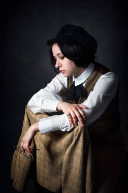 Klasik dedektif tarzı giyinmiş kadın bir Sherlock Holmes modelini somutlaştıran genç bir kadının sanatsal portresi. Sessiz bir düşünceyle oturuyor, dizine ekose bir ceket giyiyor ve bir elinde gözlük tutuyor.. 