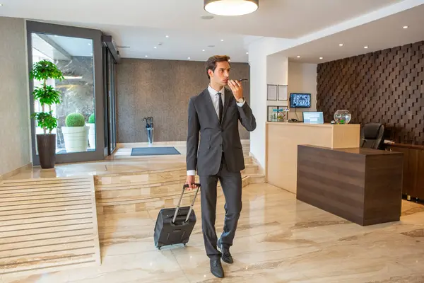 ビジネスマンはホテルのロビーでスーツケースを引っ張るとスマートフォンで会話をしています — ストック写真