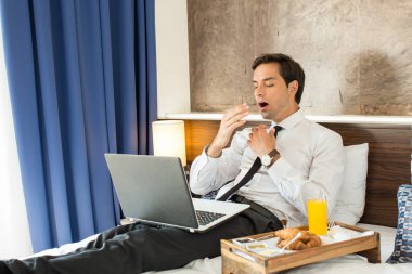 Takım elbiseli yorgun işadamı, otel yatağında bilgisayarının dizüstü bilgisayarında çalışıyor.