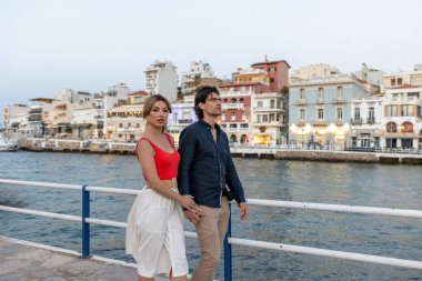 Deniz kenarında yürüyen çift, arka planda Avrupa tarzı renkli binalarla sarılıyorlar.