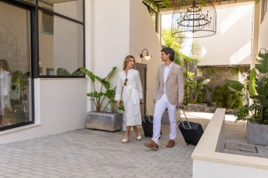 Modern bir otelin dışında yürüyen şık bir çift bavullarını çekiyor ve birbirlerine gülümsüyorlar.