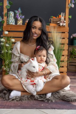 Güzel anne ve bebek, yumuşak kürklü bir halıya oturmuş, yumuşak beyaz bir tavşanı nazikçe tutuyorlar.