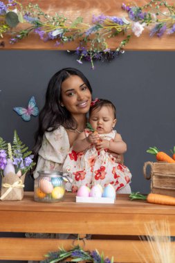 Paskalya temalı ahşap tezgah havuçla süslenmiş. Bebek bir havuç tutuyor ve çiçekli bir elbise giyiyor. Annesi yanında gülümsüyor. Etrafı renkli kelebekler ve şenlikli dekorasyonlarla çevrili.
