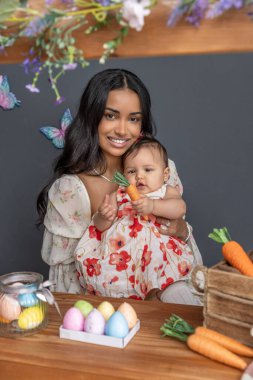 Paskalya temalı ahşap tezgah havuçla süslenmiş. Bebek bir havuç tutuyor ve çiçekli bir elbise giyiyor. Annesi yanında gülümsüyor. Etrafı renkli kelebekler ve şenlikli dekorasyonlarla çevrili.