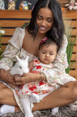 Güzel anne ve bebek, yumuşak kürklü bir halıya oturmuş, yumuşak beyaz bir tavşanı nazikçe tutuyorlar.