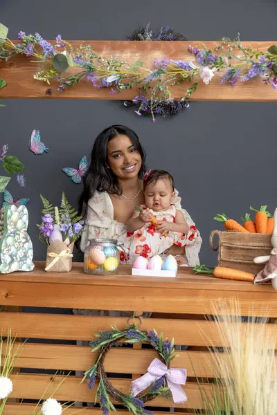 Anne ve bebek Paskalya temalı ahşap bir stantta havuç, pastel yumurta ve bahar çiçekleriyle süslenmiş.
