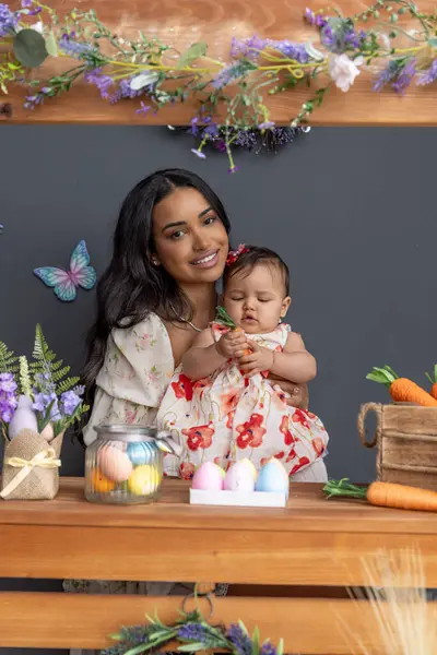 Paskalya temalı ahşap tezgah havuçla süslenmiş. Bebek bir havuç tutuyor ve çiçekli bir elbise giyiyor. Annesi yanında gülümsüyor. Etrafı renkli kelebekler ve şenlikli dekorasyonlarla çevrili.