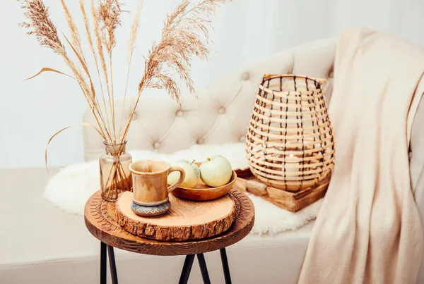 Cozycore or cottagecore concept, warm soft brown beige interior design objects. Cozy wool plaid on sofa, candle burning in wood lantern, tea cup on wood table. Warm color photo manipulation.