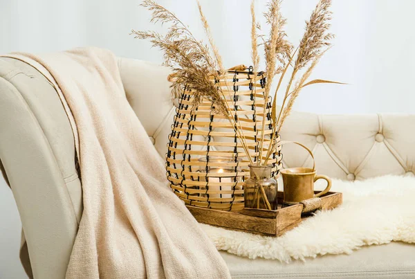 Cozycore or cottagecore concept, warm soft brown beige interior design objects. Cozy wool plaid on sofa, candle burning in wood lantern, tea cup on wood serving tray. Warm color photo manipulation.