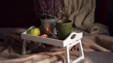 Şirin, beyaz ahşap kahvaltı tepsisi. Bacakları ev yatağında, bir fincan sıcak çay, çay mumu ve funda çiçeğiyle. Eşarp ve örgü süveter ile sonbahar düzenlemesi. İskandinav kızlık zarı yaşam tarzı.