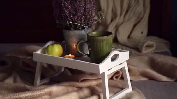 熱いお茶 お茶ライトキャンドルやヘザーの花のカップと自宅のベッドの上に足を持つかわいい白い木製の朝食トレイ スカーフとニットセーターで秋の配置を設定します 北欧の巨大ライフスタイル — ストック動画