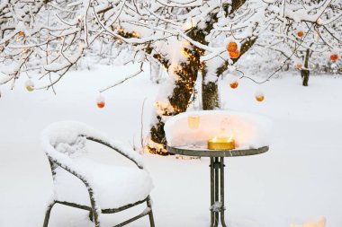 Kışın bahçedeki elma ağacını kaplayan kar, bir sürü bakır metalik Noel süsü ve ışık saçan sıcak beyaz iplikle süslenmiş. Sandalyeli masa, şarap kadehi ve açık hava mumu.