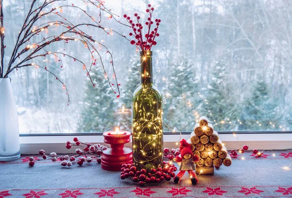圣诞装饰摆设上酒瓶 酒瓶里装满了小型的派对彩灯和用过的酒塞做成的云杉树 可爱的老式精灵雕像 后面是雪地乡村森林的窗户 — 图库照片
