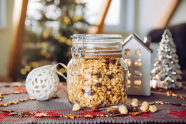 stock image Concept of homemade Christmas present, handmade roasted granola with various nuts and raisins in beautiful glass jar. Blur Christmas tree and lights on background.