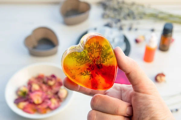 Person hand holding handmade heart soap with crystal clear gel melt and pour soap mixture. Ready to use pink and yellow soap with rose petals and lavender blossoms inside, surrounded by ingredients.