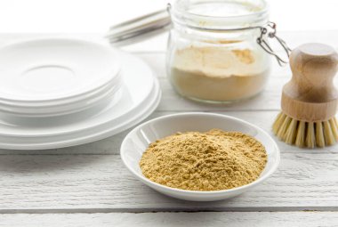 Washing the dishes with mustard powder concept. White clean plates with bowl and jar of yellow mustard powder and wooden dish washing brush and class jar container with natural washing brush.