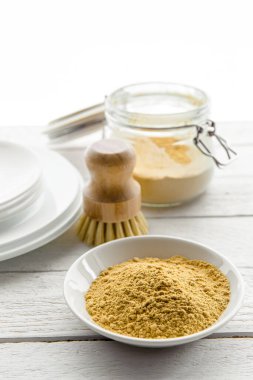 Washing the dishes with mustard powder concept. White clean plates with bowl and jar of yellow mustard powder and wooden dish washing brush and class jar container with natural washing brush.