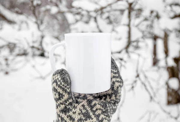 Traditional pattern mitten holding white blank coffee mug mock up template. Cozy seasonal products advertisement background. Single cup on snowy Scandinavian winter nature background. Room for text.