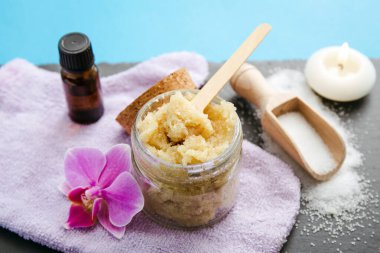 Side view of homemade sugar scrub in glass jar with ingredients for decoration, sugar on spoon and aroma oil, essential oil on blue background. Pink orchid blossom for decoration. clipart