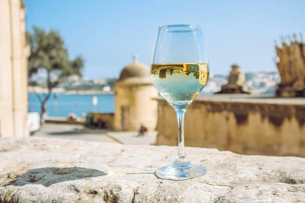 Selective focus on glass of white wine in beautiful city of Valletta in Malta. Background with blue sky and sea on sunny day. Wine tour concept.