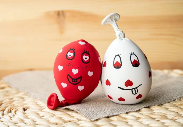Two funny homemade sensory stress balls, made of balloons and filled with flour. Red and white balls with hearts and funny drawn faces, wood background.