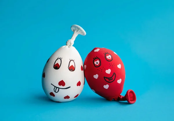 Two funny homemade sensory stress balls, made of balloons and filled with flour. Red and white balls with hearts and funny drawn faces on blue minimal background.