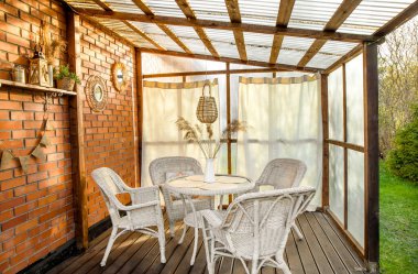 Perdeleri ve hasırları olan Boho tarzı ev terası, rattan, kamış süslemeleri. Sera plastiği ve tavandan yapılmış duvar polikarbonattır. Beyaz bahçe mobilyaları, tuğla duvardaki aynalar.