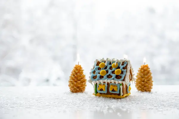 Spruce tree shape candles burning on white snowy background with winter landscape with cute gingerbread house. Lot of copy space. Winter and Christmas products background.