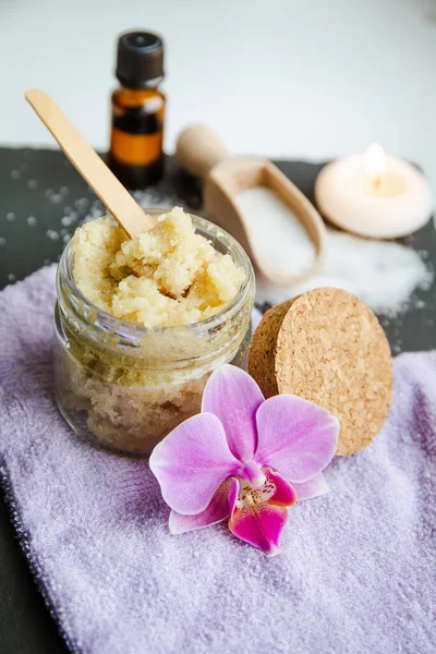 Handmade sugar body scrub in glass jar. Burning candle and pink orchid blossom for decoration on black stone white studio background.