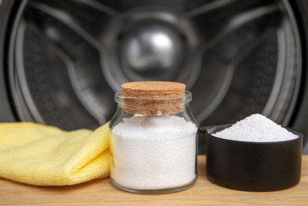 Cleaning washing machine drum with Citric acid. Measuring cup filled with citric acid and glass jar, washing cloth against washing machine drum in home indoors.