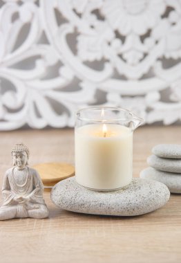 Ev yapımı masaj yağı mumu, cam bardakta, ağız ile. Bohem tarzı arka plan, oturan Buda heykeli, kompozisyonu rahatlatmak için zen taşları dizilmiş.