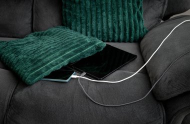 Akıllı telefon ve tablet bilgisayar oturma odasındaki yastığın altında koltukta şarj oluyor. Aşırı ısınma ve yangın tehlikesi kavramı.