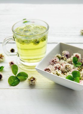 Beyaz yonca Trifolium berrak bir bardakta bitki çayına tövbe eder. İçinde ve kasesinde parıldayan çiçekler olan dumanı tüten sıcak bir içecek, arka planda çok az beyaz tahta var..