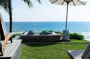 Sahile, denize, plaj şemsiyelerine, hindistan cevizi ağaçlarına ve yeşil çimenlere bakan koltuklar. mavi gökyüzü ve çimen