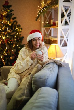 Noel Baba şapkalı heyecanlı kadın Noel ağacıyla süslenmiş oturma odasında oturmuş bir fincan çayın tadını çıkarıyor.