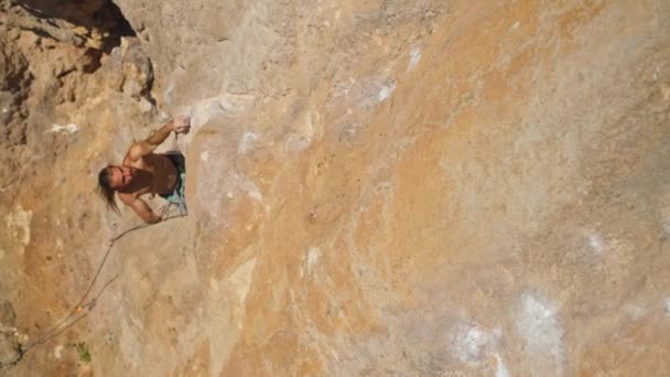 長い髪を持つ若い強いアスレチックマンロッククライマーは長い崖の上に登る 男は長いハード移動を作る非常に難しいルートを登る 極端なスポーツや屋外でのロッククライミングトレーニングの瞬間 — ストック動画