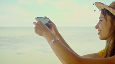 Sahilde şapkalı bir kadın dizüstü bilgisayar kullanıyor, akıllı telefon kullanıyor, internette sohbet ediyor, internette geziniyor. Teknoloji ve seyahat, tatilde online iletişim..