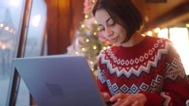Kafkasyalı beyaz kadın blogcu kırmızı şenlikli süveter giymiş kafeteryadaki kablosuz internete bağlanmış, kahve fincanı olan mutlu kadın uygulamayla sohbet ediyor. Yılbaşı iç mimarisi ve dekorasyonu