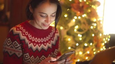 Güzel, rüya gibi esmer kadın şirin kırmızı şenlikli süveter içinde rahat kafede oturuyor telefon kullanıyor, mesaj yazıyor, tebrik ediyor ya da sosyal ağlara bakıyor. Noel süsleri, bayram havası.
