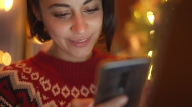 Arka planda ışıkları kullanarak, telefon ederek, mesajlaşarak, tebrik yazarak, sosyal ağları arayarak, sevimli kırmızı şenlikli süveter giyen hayalperest bir kadın. Noel süsleri, bayram havası.