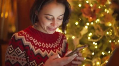 closeup cute smiling woman in red festive sweater on background of lights on christmas tree using phone, messaging, reading and writing congratulations, looking social networks. holyday mood