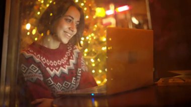 festive weared woman sits in decorated cafe close to window on Christmas Eve using laptop. girl dreamily looking at window. festive mood, making wishes, dreams concept