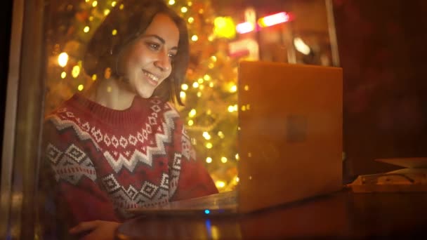 圣诞前夕 穿着节日服装的女人坐在靠近窗户的装饰咖啡店里 使用笔记本电脑 女孩梦游般地看着窗户 喜庆的心情 梦想的概念 — 图库视频影像
